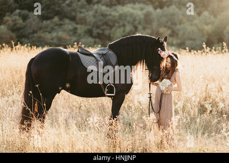 Caucasian female wearing a beige flowy dress and a flower crown, on a yellow grass field with her black horse Stock Photo