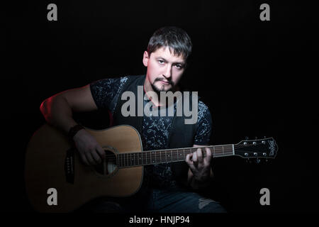 Portrait of handsome bearded man sitting in a chair with an acoustic guitar in hand, isolated on a black background Stock Photo