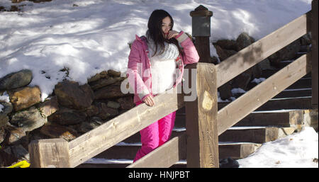 Adorable asian girl standing on wooden stairs in mountain snowy resort. She wearing snowboarder clothes. Stock Photo