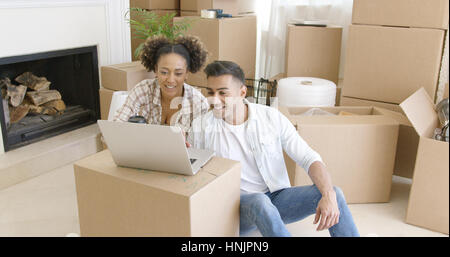 Happy mixed race couple sitting on the floor among unpacked carton boxes and using laptop computer to plan the future in their new apartment they just Stock Photo