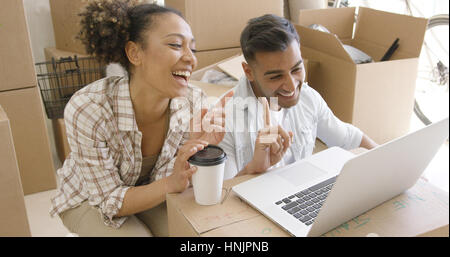 Happy mixed race couple discuss while using laptop in their new apartment. They have unpacked boxes being their back. Smiling and planning the future. Stock Photo