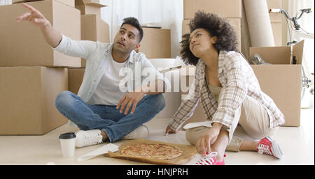 Young couple eating pizza on the floor in front of a stack of brown packing boxes as they move house and chatting  pointing to something off screen. Stock Photo
