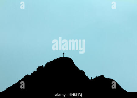 Black silhouette of mountain peak with summit cross. Blue background. Stock Photo