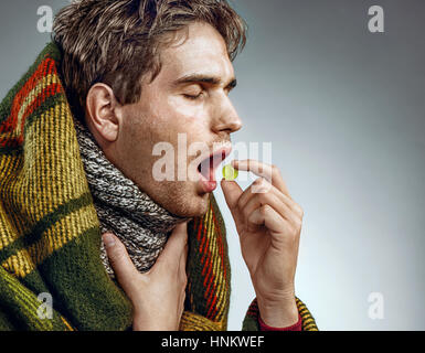 Man with open mouth take pill for throat. Photo of sick man wrapped in blanket on grey background Stock Photo