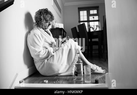 Depressed woman in dressing gown at home during daytime heavily drinking and taking tablets to self medicate phoning for help - Posed by model Stock Photo