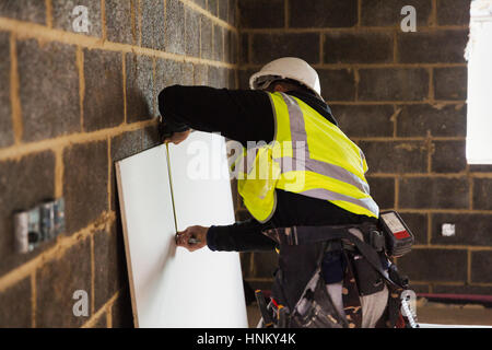 A builder in a hard hat wearing a tool belt, measuring a piece of wood. Stock Photo