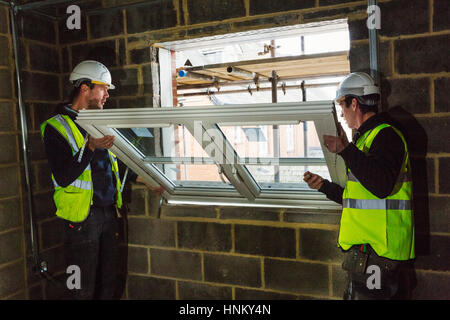 Two builders in hard hats and high visibility jacket fitting a window frame. Stock Photo