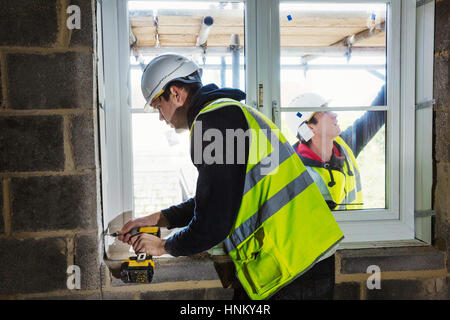 A workman on a construction site, builder in hard hat using an electric drill on a window sill. Stock Photo