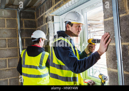 Two workmen on a construction site, builder in hard hat using an electric drill on a window frame. Stock Photo