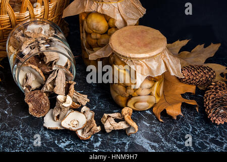 Two glass jars with wild mushrooms on black marble background. One jar with dry mushrooms and one with pickled mushrooms. Several dry white wild mushr Stock Photo