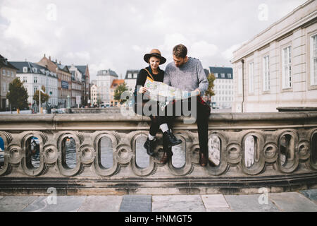 Young couple looking at a navigation map sitting on a river bridge. Tourists exploring the area on cloudy day. Stock Photo