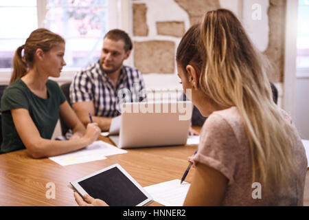 Young business woman with her hair tied back checking her notes while holding a tablet and a man and woman sit in the background Stock Photo