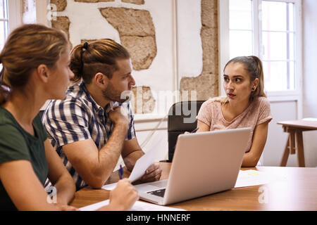 Three young business professionals in a meeting using a laptop and notes sitting at a table and their hair tied back Stock Photo