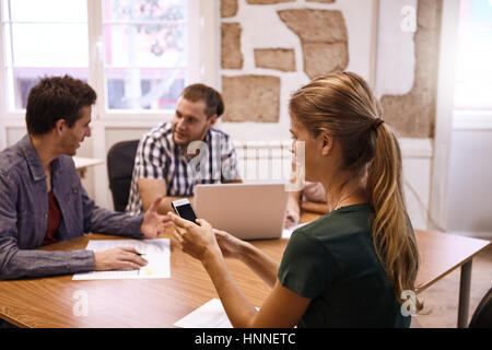 Young professional female sending message on the phone, with two young male professionals in the background in a meeting Stock Photo