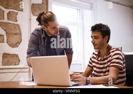 Young businessman being instructed by a male colleague while looking at his laptop on the desk in front of him Stock Photo