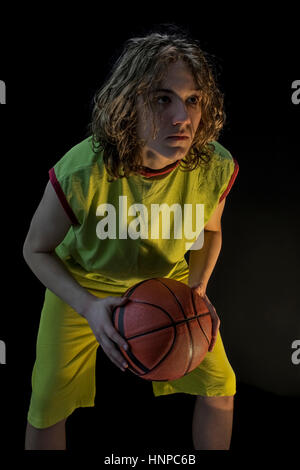 Young boy with long blond hair wearing a green jersey in a squatting position and holding a basketball focusing on the making the shot. Stock Photo