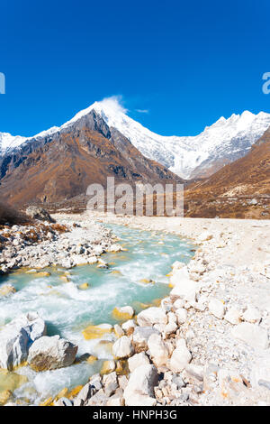 Rocky barren terrain landscape lines a fast flowing glacier water river with Himalayan mountain range, Langtang Lirung peak, in background at high alt