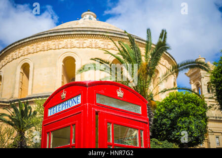 Mosta, Malta - Traditional British red telephone box with palm trees and the famous Mosta Dome at background with blue sky and clouds Stock Photo