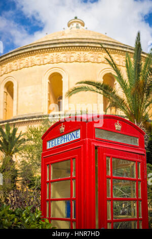 Mosta, Malta - Traditional British red telephone box with palm trees and the famous Mosta Dome at background with blue sky and clouds Stock Photo