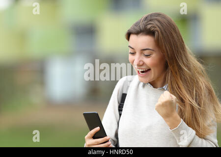 Excited student reading good news on line in a smart phone in the street with the university building in the background Stock Photo