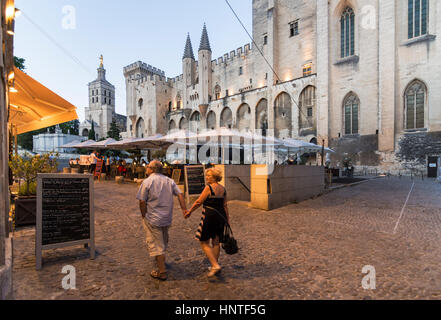 A couple walk past cafes and restaurants in Palace Square, overlooked by the Palace of the Popes in the evening, Avignon, France Stock Photo