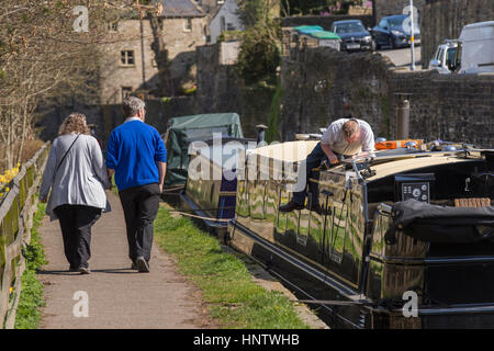 Couple walking past a man working aboard a moored canal boat, fixing a solar panal to the roof - Leeds Liverpool Canal, Skipton, North Yorkshire, GB. Stock Photo