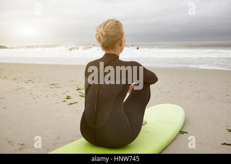 Female surfer sitting on surfboard looking out from Rockaway Beach, New York, USA Stock Photo