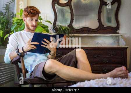 Young man with red hair, sitting in chair, reading book Stock Photo