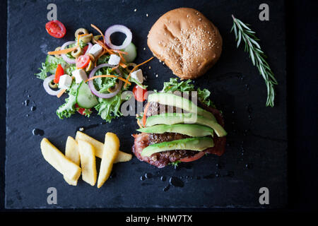 Overhead view of hamburger with avocado, side salad and chips on slate Stock Photo