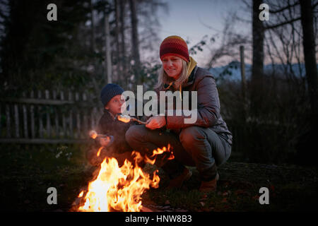 Mature woman and son toasting marshmallows on garden campfire at dusk Stock Photo