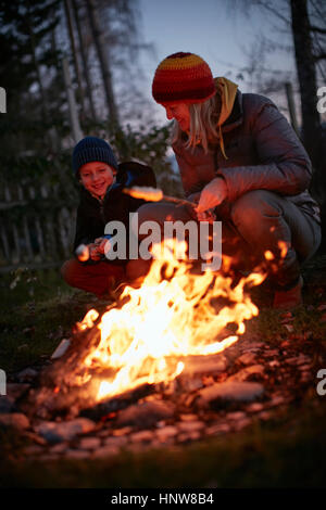 Mature woman and son toasting marshmallows on garden campfire at dusk Stock Photo