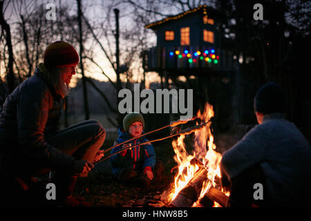 Mature woman and two sons toasting marshmallows on campfire at night Stock Photo
