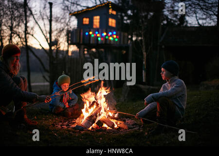 Mature woman and two sons toasting marshmallows on campfire at night Stock Photo