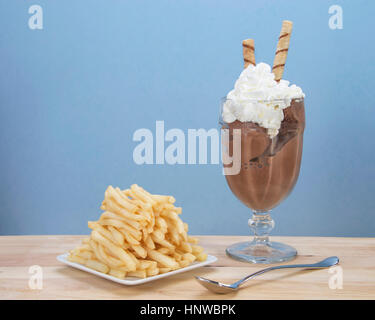 Chocolate ice cream in glass cup with whipped cream and square white plate of stacked french fries on light wood table. Food fad or new trend. Stock Photo