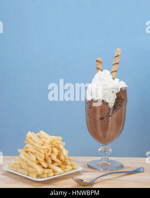 Chocolate ice cream in glass cup with whipped cream and square white plate of stacked french fries on light wood table. Food fad or new trend. Stock Photo