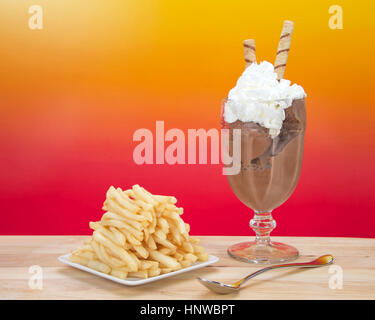 Chocolate ice cream in glass cup with whipped cream and square white plate of stacked french fries on a light wood table. Food fad or new trend. Stock Photo