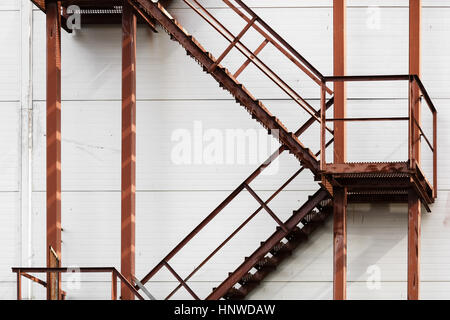 Old rusty metal ladder against a white wall background. Metal steps with handrails and staircases covered with rust. Facade of industrial buildings. Stock Photo