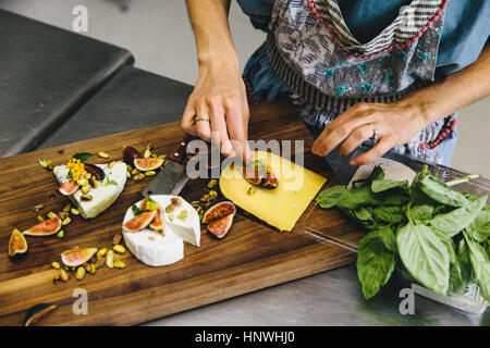 Woman preparing variety cheese plate with figs, nuts, pistachios, basil on wooden chopping board Stock Photo