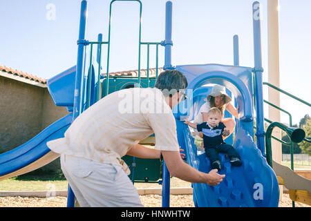Family at playground, young son sliding down slide Stock Photo
