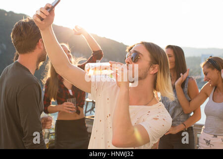 Young man taking selfie drinking beer at waterfront roof terrace party, Budapest, Hungary Stock Photo