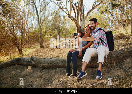 Mid adult couple hiking, sitting on log taking smartphone selfie Stock Photo