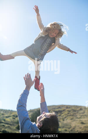 Girl being thrown mid air by father against sunlit blue sky Stock Photo