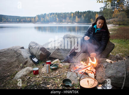 Woman Sitting Near Bonfire On Lakeshore During Camping Stock Photo