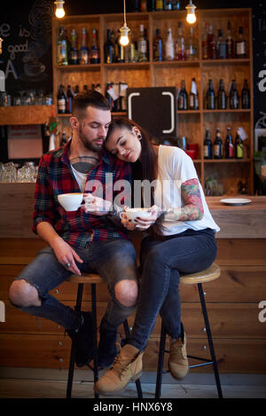Couple sitting side by side on stools in cafe, head on shoulder Stock Photo
