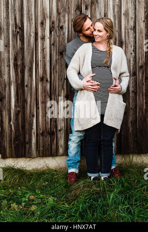 Man kissing pregnant girlfriend on cheek by wooden fence Stock Photo