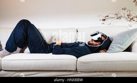 Mid adult man lying back on sofa watching virtual reality headset Stock Photo
