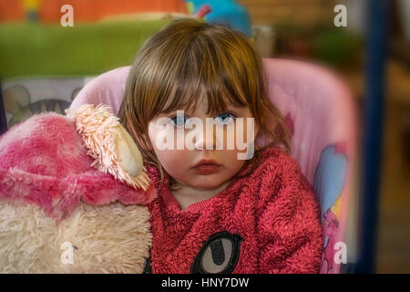 Blond baby girl with vibrant blue eyes wearing a pink fur sweater, looking innocently in the camera and holding a furry teddy bear. Stock Photo