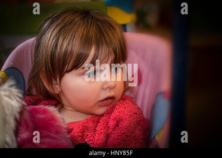 Blond baby girl with vibrant blue eyes in a pink fur sweater sitting on a swing and looking to the side. Stock Photo