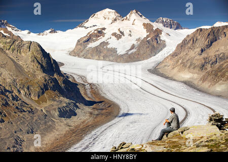 Young man sitting and enjoying a majestic view to Aletsch glacier, the largest gracier in Alps and UNESCO herritage from Eggishorn, Valais, Switzerlan Stock Photo