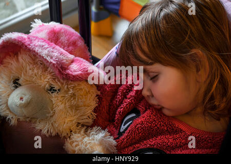 Blond baby girl wearing a pink fur sweater asleep with a furry teddy bear in her hands. Stock Photo